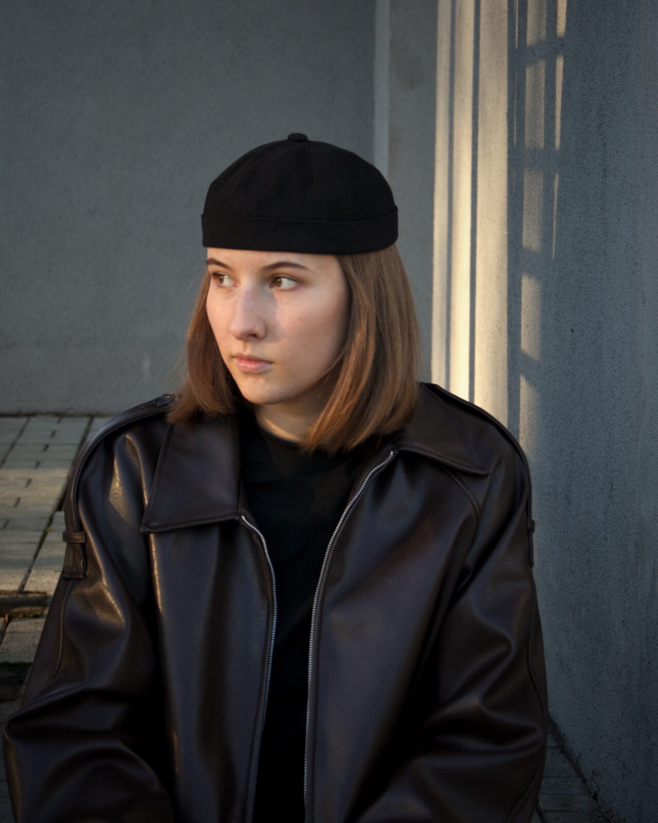 Sofia Lopato — портрет: чёрная кожаная куртка и чёрная шапка-докер, вечерний свет из-за бетонной колонны
